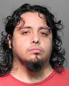A man with medium-length, curly black hair and a goatee. He has a small tattoo near his left eye. He is wearing a red shirt and has a neutral expression on his face.