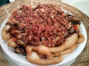 A plate of funnel cake topped with a generous layer of crumbled bacon and drizzled with what appears to be chocolate or syrup. The dessert is set in a woven basket.