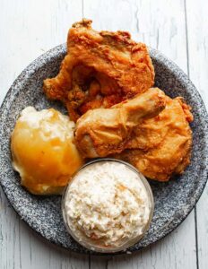 A gray speckled dinner plate holding two pieces of fried chicken, a scoop of mashed potatoes with brown gravy, and a small glass bowl of creamy coleslaw. The fried chicken pieces are golden-brown and crispy-looking, taking up the upper and right side of the plate. The mashed potatoes with gravy sit on the left side. The coleslaw in the glass bowl is placed near the bottom centre of the plate.
